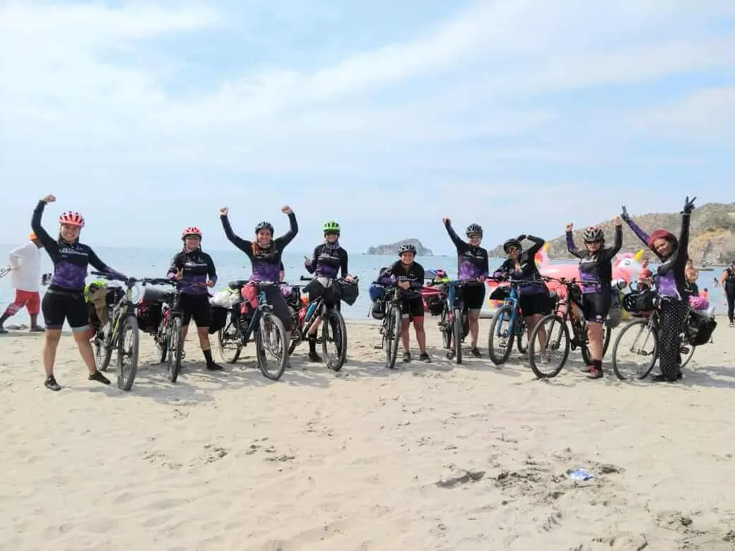 Grupo de mujeres ciclistas con los brazos en alto junto a sus bicicletas cargadas en una playa