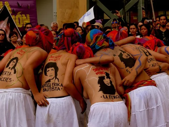 Mujeres abrazadas de espaldas con faldas blancas y rostros pintados en la espalda, en una manifestación