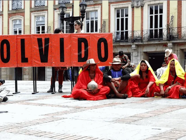 Mujeres con mantos rojos sentadas en una plaza frente a una pancarta con la palabra «OLVIDO»
