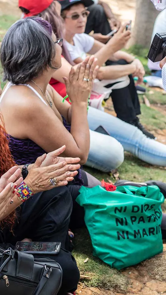 Mujeres sentadas en el pasto aplauden junto a un bolso verde con la frase «Ni dios ni patria ni marido»