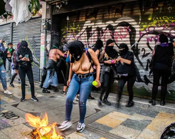 Mujeres con el rostro cubierto y una de ellas con el torso desnudo, junto a una hoguera frente a una cortina metálica con grafitis