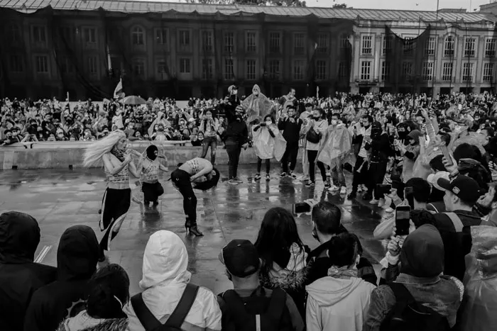 Multitud bajo la lluvia frente al Capitolio, con jóvenes encapuchados en primer plano, en blanco y negro