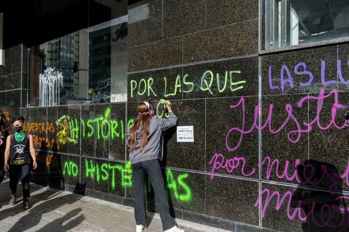 Muro de granito con consignas pintadas en verde y morado, como «Por las que…» y «Justicia», mientras una persona pinta