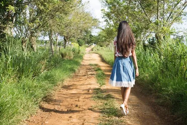 Niña de espaldas con vestido azul camina descalza por un camino de tierra entre árboles y pasto