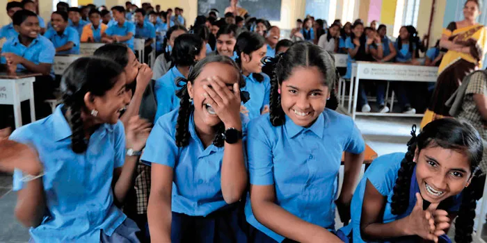Niñas con uniforme azul se ríen sentadas en un salón de clases lleno de estudiantes