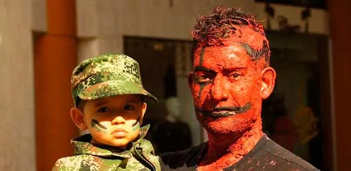 Niño con gorra y pintura de camuflaje junto a un hombre con el rostro cubierto de pintura roja