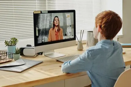 Niño de espaldas sentado ante un computador donde una mujer aparece en videollamada