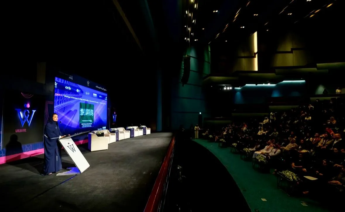 Oradora ante un atril en un escenario con un panel y un auditorio lleno de público