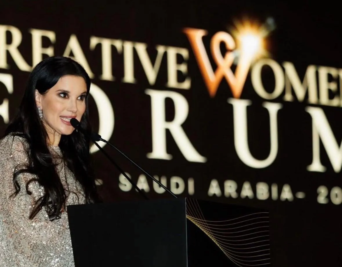 Mujer de cabello largo habla ante un micrófono bajo el letrero «Creative Women Forum Saudi Arabia»