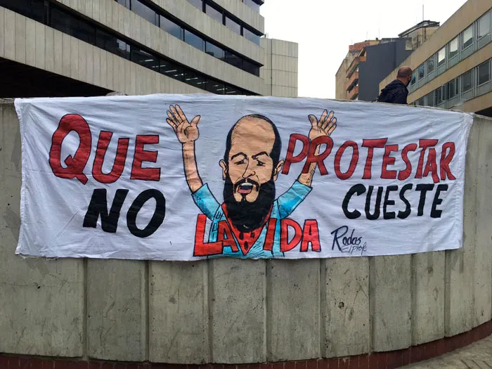 Pancarta «Que protestar no cueste la vida» colgada de un muro de concreto, con un dibujo de un hombre
