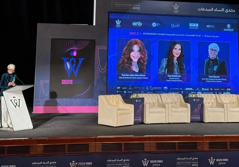 Panel en un escenario con una pantalla que muestra a tres ponentes y una oradora en el atril