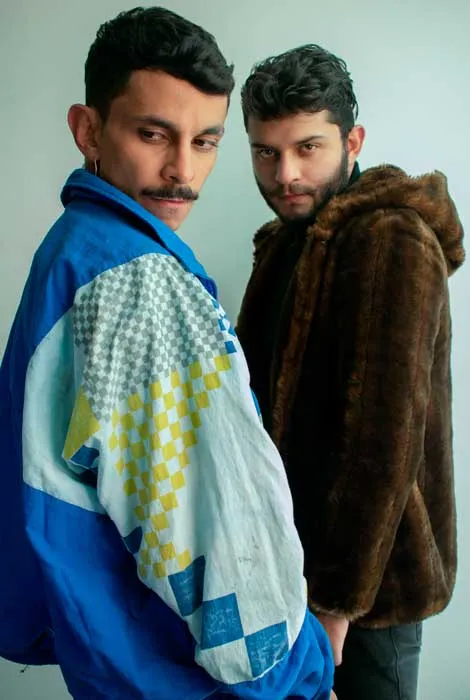Dos hombres jóvenes posan lado a lado, uno con chaqueta deportiva azul y otro con abrigo de pelo marrón