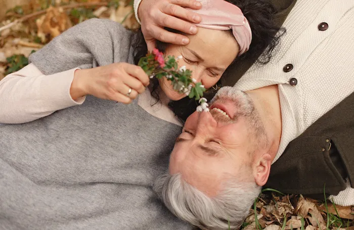 Pareja de personas mayores recostada en el suelo, sonriendo con los ojos cerrados y una flor entre sus rostros