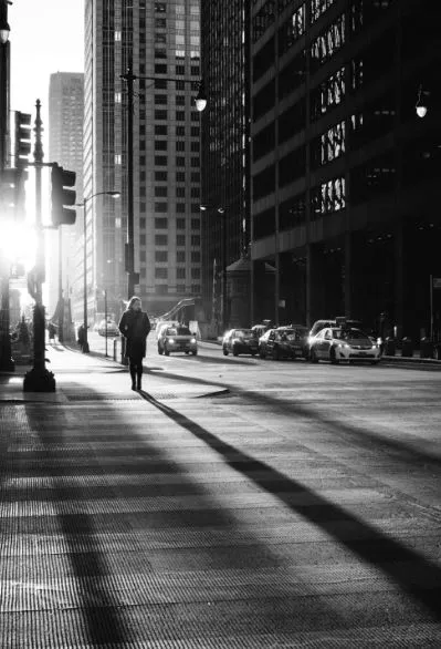 Persona caminando sola por una calle de ciudad con largas sombras entre edificios, en blanco y negro