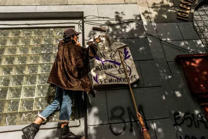 Persona con capa marrón sostiene un cartel feminista junto a un muro pintado con «8M»