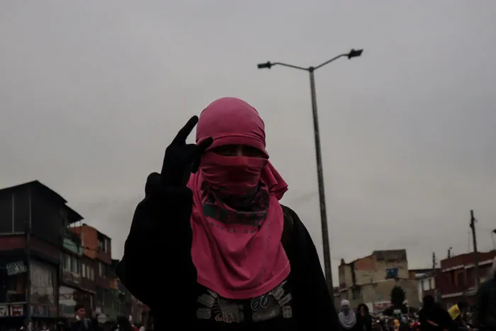 Persona con pasamontañas rosado hace un gesto con la mano bajo un cielo gris en una calle de Bogotá
