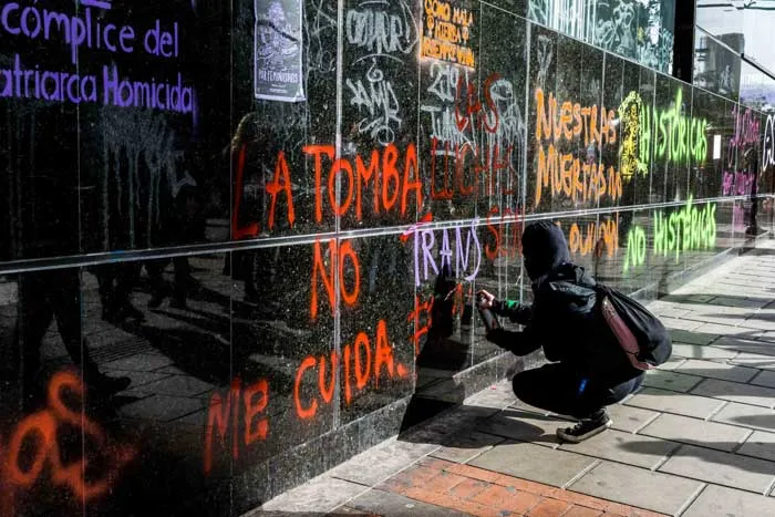 Persona agachada pinta consignas sobre un muro negro, entre ellas «cómplice del patriarcado homicida», durante la marcha del 8 de marzo