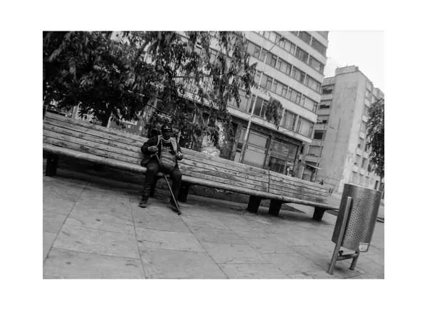 Persona sentada sola en una banca de una plaza junto a edificios, en blanco y negro