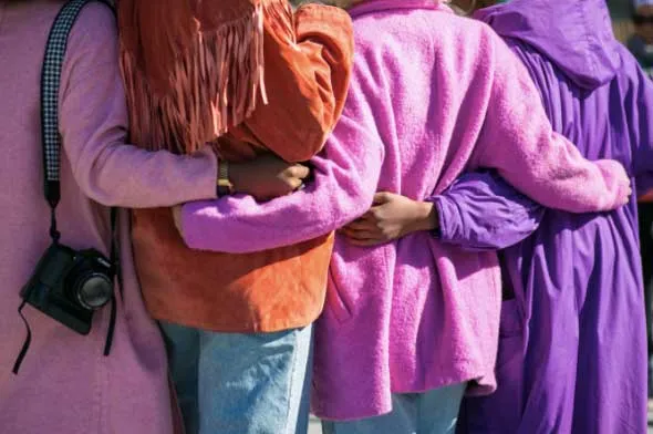 Tres personas de espaldas con abrigos de colores se abrazan por los hombros, una con cámara colgada