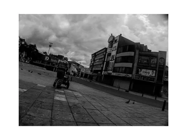 Plaza amplia y casi vacía con un vendedor en un triciclo y edificios al fondo bajo un cielo nublado, en blanco y negro