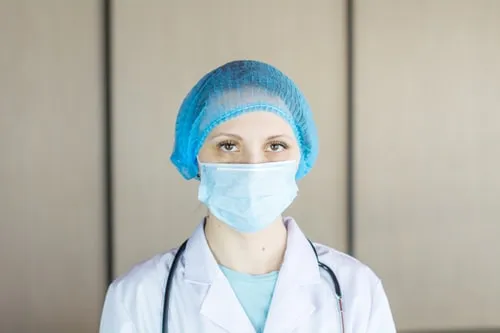 Profesional de la salud con bata blanca, gorro azul, mascarilla y estetoscopio mira a la cámara