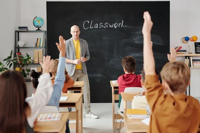 Profesor frente a una pizarra con la palabra «Classwork» y alumnos que levantan la mano en un aula