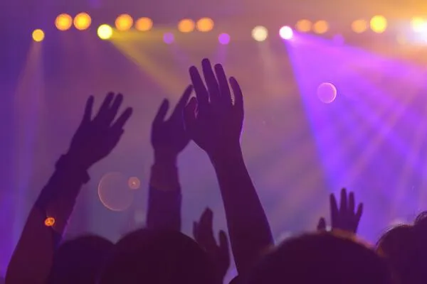 Manos en alto del público ante un escenario con luces moradas y naranjas en un concierto