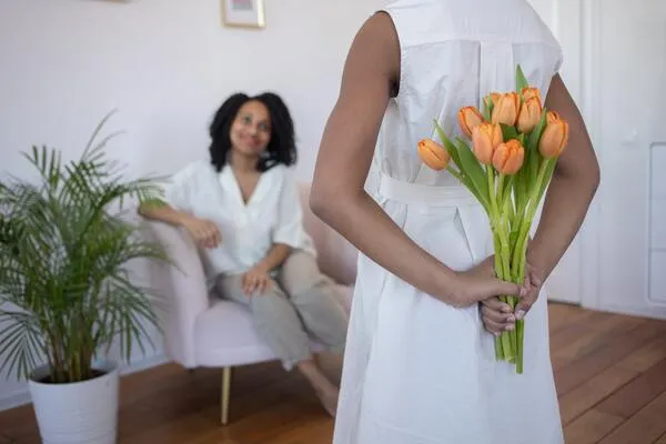 Persona de espaldas con vestido blanco sostiene un ramo de tulipanes naranjas mientras una mujer sonríe sentada al fondo