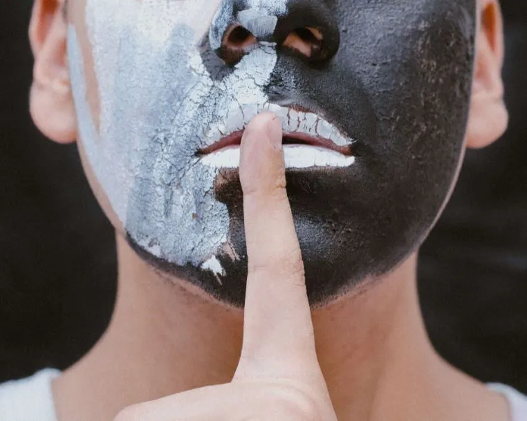 Rostro pintado de negro y blanco agrietado con un dedo sobre los labios pidiendo silencio