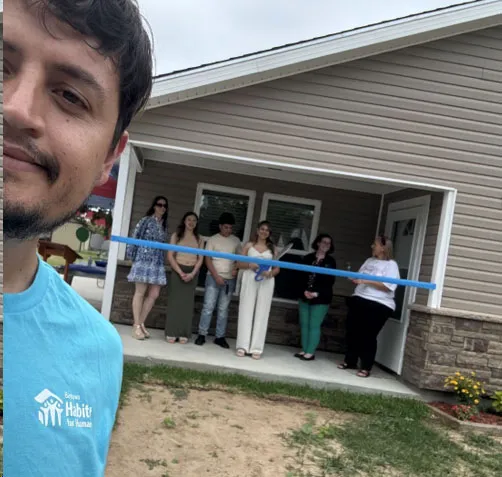 Selfi de un hombre con camiseta de Habitat for Humanity con un grupo de personas frente a una casa con una cinta azul