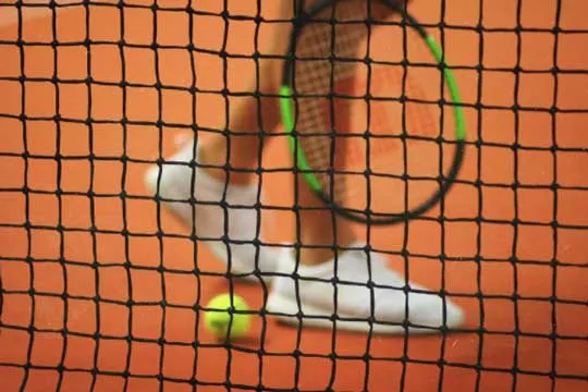 Piernas y raqueta de una tenista vistas a través de la red sobre cancha de arcilla, con una pelota junto a sus zapatillas