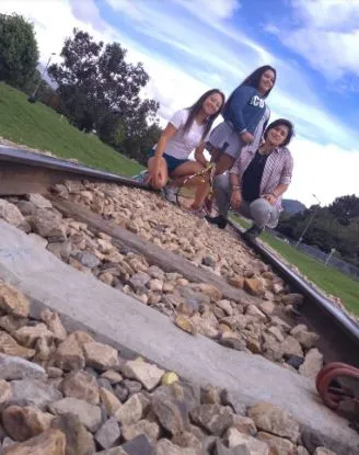 Tres mujeres jóvenes sentadas junto a una vía de tren rodeada de piedras, bajo un cielo con nubes